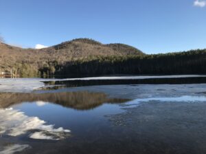 A forested hill stretches across a blue sky. It sits behind a large clear lake covered with melting ice.